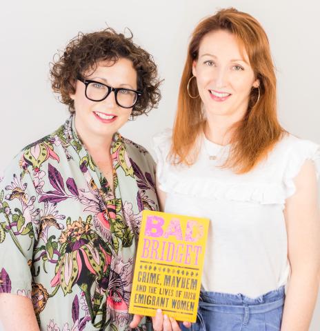 Two women stand side-by-side holding up the book they have written, 'Bad Bridget'. One woman wears glasses and has short curly hair, the other woman has long red hair.