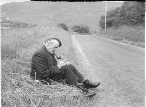 black and white photograph of a man smoking a pipe and sitting at the side of the road. He is writing in a notebook