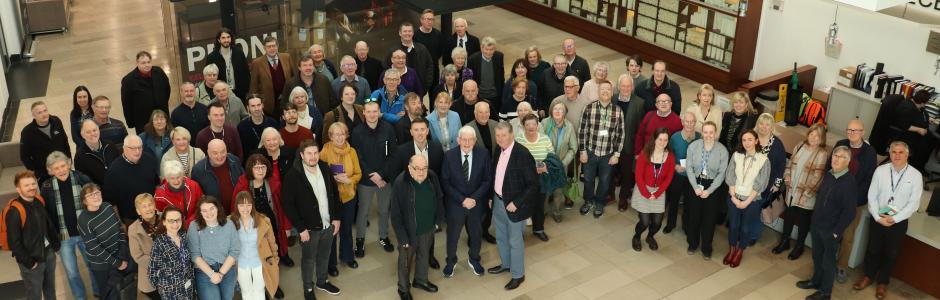 Birdseye view of a group of about 50 to 60 men and women looking up at the camera. They stand in a large atrium with a visible staircase behind them.