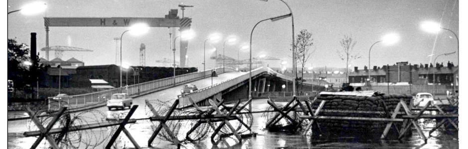 Black and white image of a bridge with a crane in the background and an army barricade in the foreground.