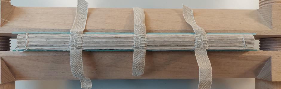 Photograph of the side of a medium size volume wedged in a large wooden vice. The side of the volume has straps fastened in three parallel lines with thread sewen over the top. The front and back cover of the volume is pale blue and just visible on the top and bottom.