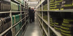 Image taken in between two long rows of shelves containing film reels with two staff members standing in between.