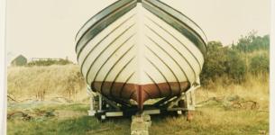 Front-view photograph of a white boat with green trim, with grass and bushes in the backdrop.