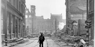 Soldier in uniform with a bayonet walking on an empty street with damaged buildings and debris in the background.