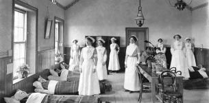 Black and white image of eight women in nurse uniforms attending to beds of injured men in a large room with a table and stools.
