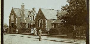 Sepia tone photograph of a school building behind a fence on a street with children dressed for school.