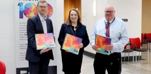 Two men and a woman in between standing in an office building holding copies of a multicoloured book.
