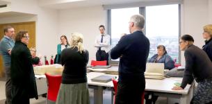 Group of ten people around a table with archival volumes and boxes. 2 of the group are sitting in red chairs and a large open window is in the background.
