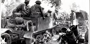 Black and white photograph of 6 army soldiers sitting in large open-back vehicles on a grass path with trees in the backgroud. Large amounts of army equipment including helmets are visible.
