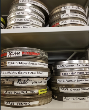 Two shelves with stacks of silver film reels and handwritten labels on the front of them.