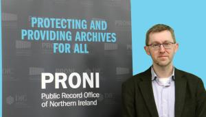 Image of a man with short brown hair and glasses standing to the right of a poster with the words 'Protecting and Providing Archives For All' in light blue text, followed by 'PRONI - Public Record Office of Northern Ireland' in white text. The wall in the background is also light blue.