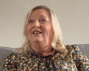 Headshot of a middle aged woman with medium-length blond hair and a colourful floral blouse talking on a couch. 