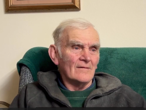 Headshot of an older man with a green top and grey hoodie sitting on a green couch against a light yellow wall and the bottom of a frame in the background.