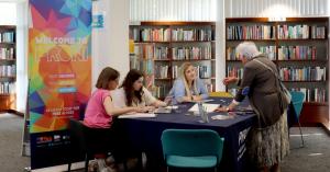 PRONI staff members and library visitors talking around a table. In the background are shelves of books and a popup banner that reads 'Welcome to PRONI'