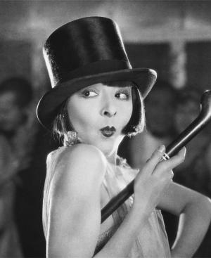 black and white photograph of a woman with short bobbed hair, wearing a top hat and holding a cane under her arm. She is looking behind her with her lips puckered