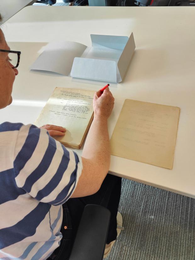 Woman with glasses and horizontal striped t-shirt sitting at a table writing page numbers on an original document. A four flap folder sits open on the table above the document.