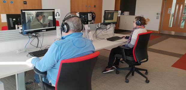 Man and woman wearing headsets and watching video clips on the computer while sitting in office chairs.