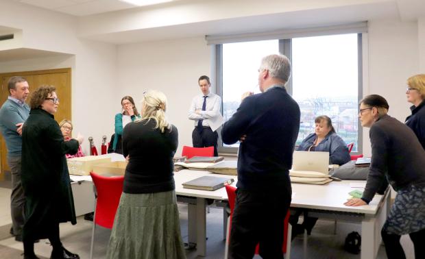 Group of ten people around a table with archival volumes and boxes. 2 of the group are sitting in red chairs and a large open window is in the background.