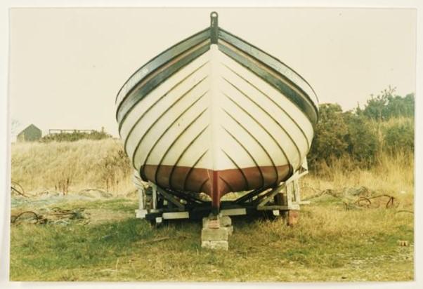 Front-view photograph of a white boat with green trim, with grass and bushes in the backdrop.