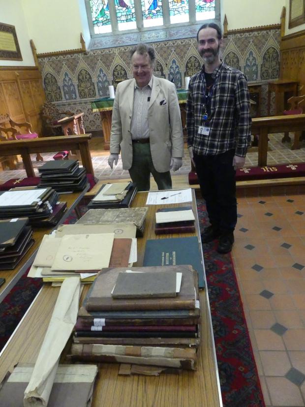 Two men standing in a church by a large table with many books and documents. 