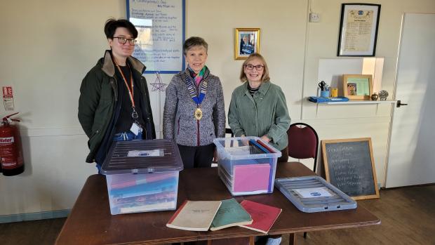 Three people standing behind a wooden table with two boxes on it. One of the boxes is open and there are three volumes on the table.