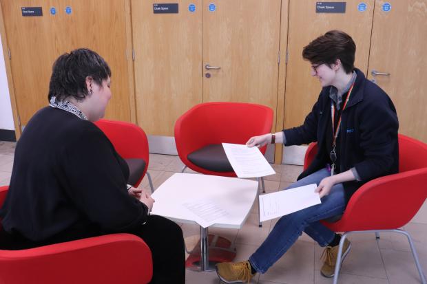 Two people sitting at a small table with four red chairs around it, looking at papers together.