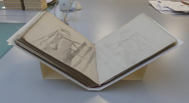 Large book open in a v shape with a support under the spine. The open pages display line drawings.
