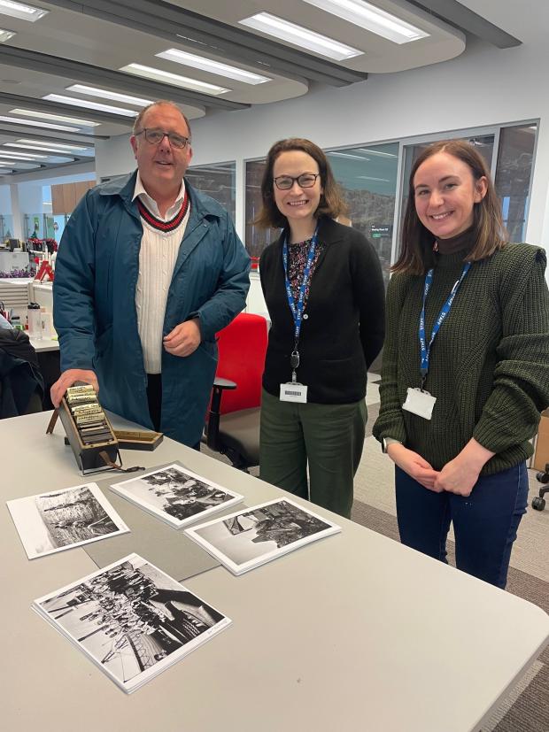 One man and two women with lanyards standing by a large table with black and white photographs displayed. 