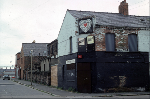 Image of a street corner showing a row of brick buildings. The building in the foreground has multiple signs for alcohol brands and a street sign reading "Grovefield Street". 