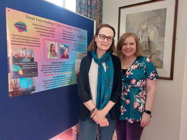 Two women standing next to a large colourful poster with text and images.