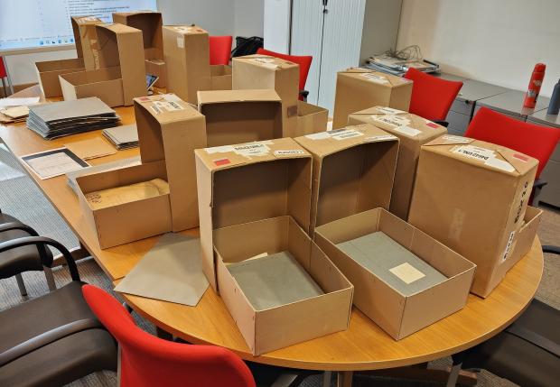 Around eight archival boxes with open lids on a large table, with grey files visible inside.
