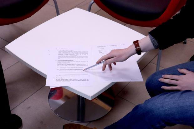 Small white table with papers and 2 red and black chairs visible. A staff member points to sections of the papers with a pencil.
