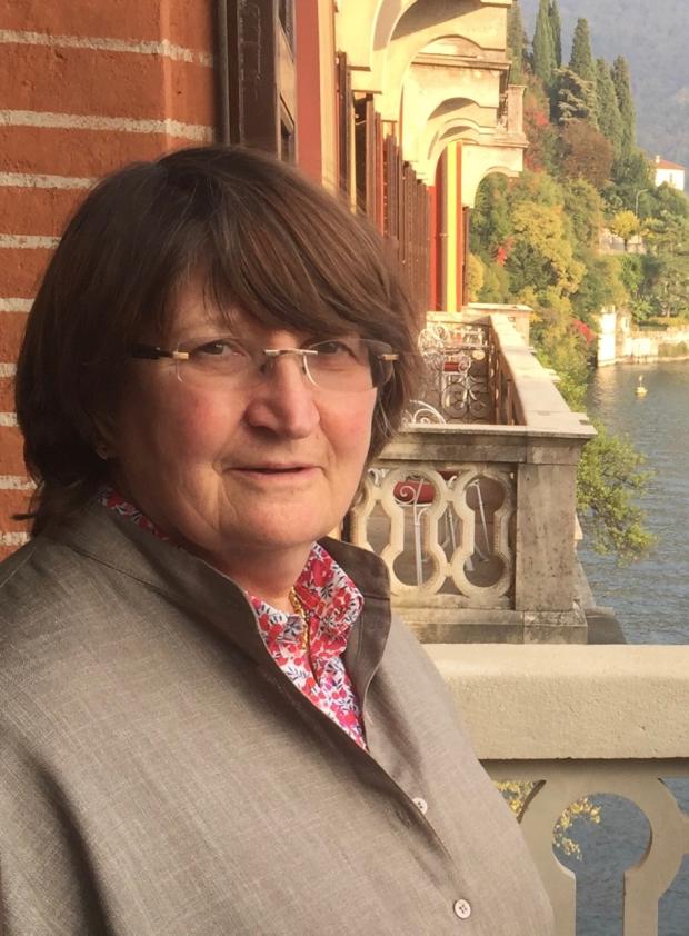 Side angle photo of a woman with chestnut brown hair standing on a balcony of a red brick building.