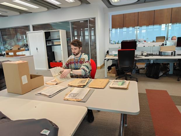 Young man with a colourful jumper sitting at a large table in an office sorting archival papers next to an open archival box.