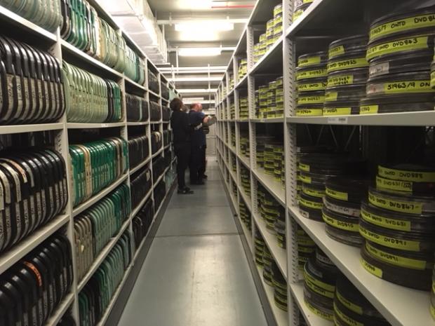 Image taken in between two long rows of shelves containing film reels with two staff members standing in between.