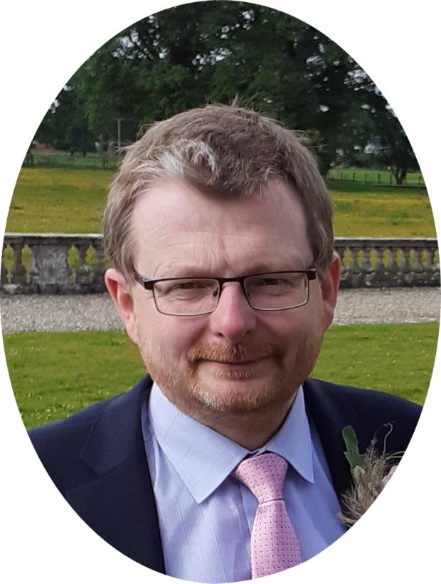 Headshot of a man with light brown hair, wearing a suit and pink tie with a floral button hole.