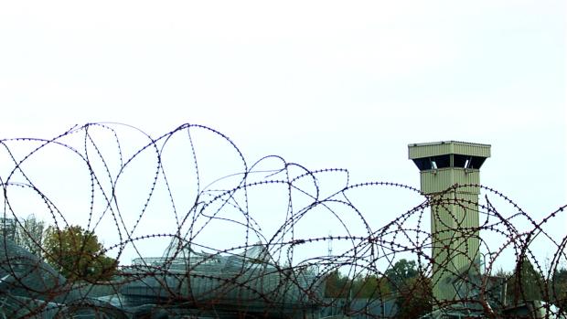 Image of a white watchtower visible over a row of barbed wire. Trees and other shrubs are visible in the background.