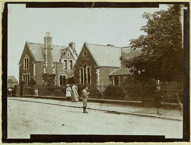 Sepia tone photograph of a school building behind a fence on a street with children dressed for school.