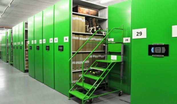 Large store with a long row of bright green bays and electrical panels on the front. Visible in the four ground are shelves with archival boxes and a large green step ladder.