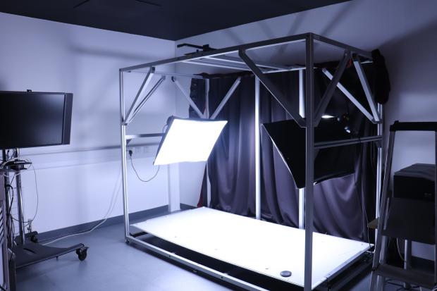 Dark room featuring a large photography set up of two large light panels over a large rectangular platform. A mobile television set is visible in the left hand side.