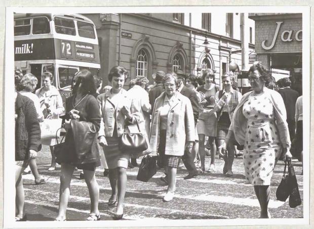 Black and white photograph of multiple women walking in different directions across a busy high street, with a  bus in the background.