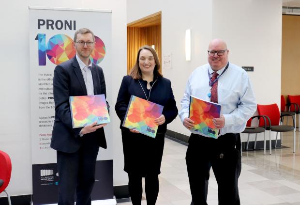 Two men and a woman in between standing in an office building holding copies of a multicoloured book.