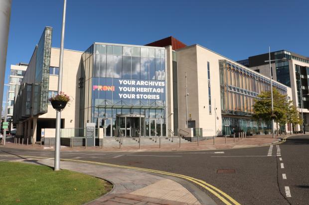 Large building in the sun with a glass front with a road in front that bends round to the left.