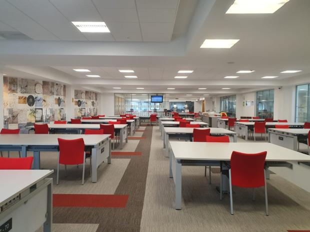 Image of a large room with multiple long tables and red chairs on either side. A tv screen is visible at the far end.