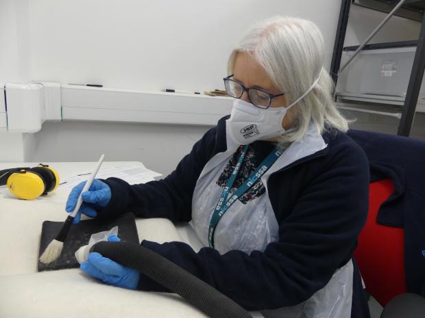Woman sitting at a white desk with protective equipment on (mask, gloves, apron) cleaning a document with large tools.