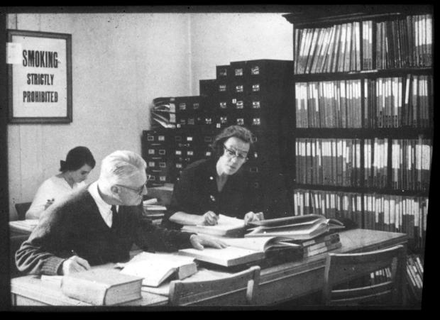 Black and white photograph of two women and one man sitting at large oak desks with open books. In the background is a large bookcase, large filing cabinets and a sign reading 'smoking strictly prohibited'.