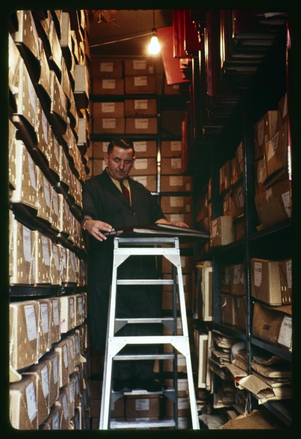 Man in a suit and tie standing on a step ladder in a small room with shelves stacked with archival boxes.