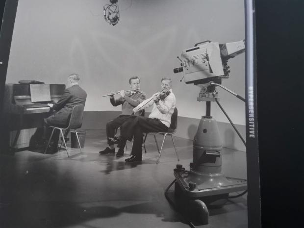 Black and white image of three men in a studio playing instruments, piano, fiddle and a flute, with a large camera next to them and a microphone overhead.