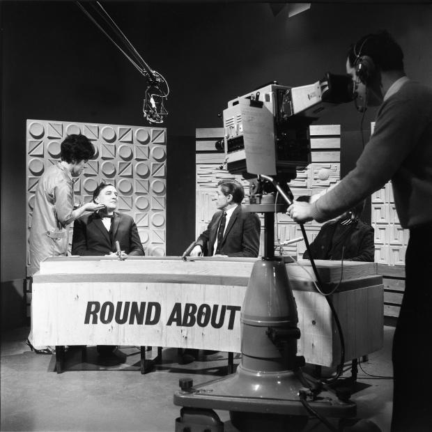 Black and white image of two men at a news style desk, one of whom is being touched up by a make up artist. The room also features a man looking into a large wheelable camera in the foreground.