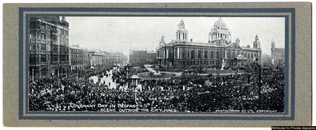 Black and white photograph of a very large crowed by a large town hall and gardens.